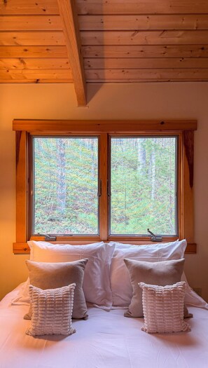 A bedroom with a large window, wooden ceiling, and a bed with white bedding and two decorative pillows.