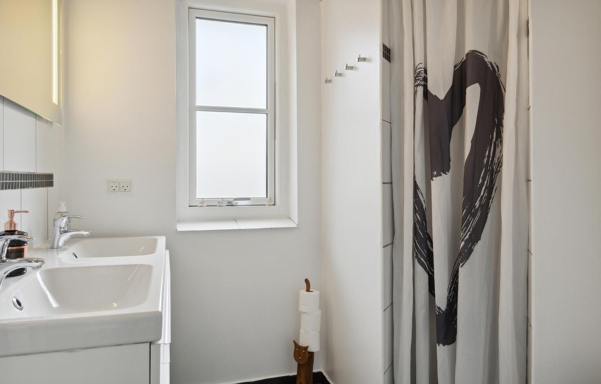 Une salle de bain avec un lavabo blanc, un miroir, une fenêtre et un rideau de douche orné d’un motif abstrait noir.