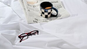 A bed with white sheets, a checkered mug, a newspaper, and a pair of glasses. - Dye House (Providence)