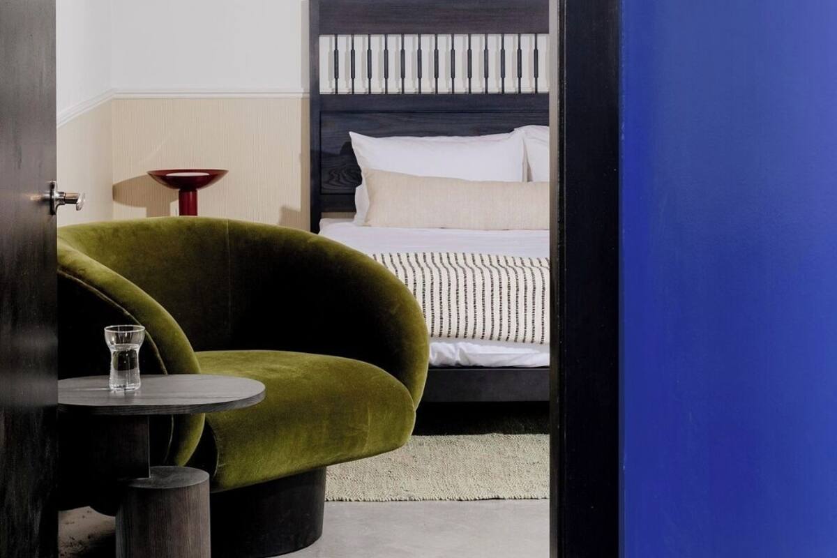 A room with a bed, a green armchair, a small round table with a glass, and a red lamp.