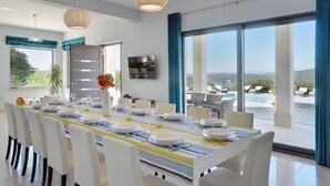 A dining area with a long table set for six, large windows with a view of a pool and mountains, and two hanging light fixtures.