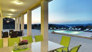 A covered outdoor seating area with a pool and lounge chairs, overlooking a mountainous landscape.