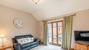 Een woonkamer met een zwarte leren bank, een houten tv-meubel, een grote flatscreen-televisie, een wandklok en een glazen schuifdeur die uitkomt op een terras.