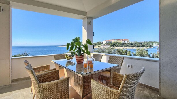 A balcony with wicker chairs and a glass table, overlooking a coastal view with a building in the background.