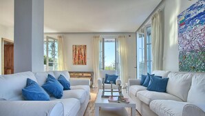 A spacious living room with two white sofas and a coffee table, large windows with curtains, and a colorful abstract painting on the wall.