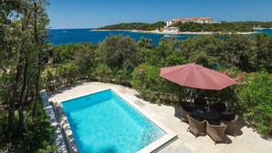 A swimming pool with a view of the sea, surrounded by greenery and a building in the background.