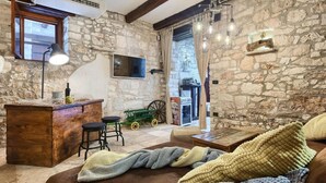 Une pièce rustique aux murs de pierre, avec un bar en bois, des tabourets de bar, un téléviseur et un coin salon confortable avec un canapé et des couvertures.