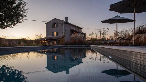 Una piscina con reflejo claro, una casa de techo inclinado y mobiliario exterior bajo sombrillas.