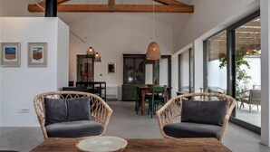 Una habitación amplia con techo alto, vigas de madera, sillas de mimbre con cojines, una mesa de madera con un cuenco y vistas a un área exterior con sillas a través de amplias ventanas.