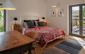 Ein Schlafzimmer mit Bett, einem Holztisch, einem Stuhl, einem Nachttisch und einer Lampe. Es gibt ein Fenster mit Blick auf Grünflächen und einen Balkon mit Gartenmöbeln.