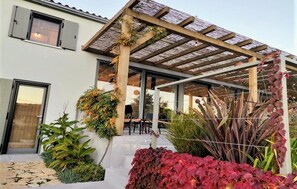 Ein modernes Haus mit überdachter Terrasse, Sitzgelegenheiten im Freien und verschiedenen Pflanzen.