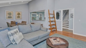 A living room with a grey sofa, a coffee table with a newspaper, a wooden ladder, and a staircase leading to another level. - Hilton Lake Getaway w/ Hot Tub & Two Fire Pits (Chapin)