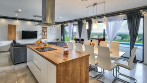 A modern kitchen with a central island, dining area with white chairs, and a large window overlooking a pool.