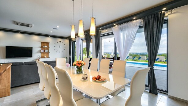 A modern dining area with a white table and chairs, a clock, and a view of a swimming pool.