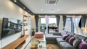 A modern living room with a flat-screen TV, a sofa, a glass coffee table, and a dining area visible through the glass doors.