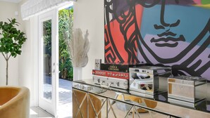 Een modern interieur met een glazen tafel waarop bordspellen liggen, een groot abstract schilderij en uitzicht op groen buiten.
