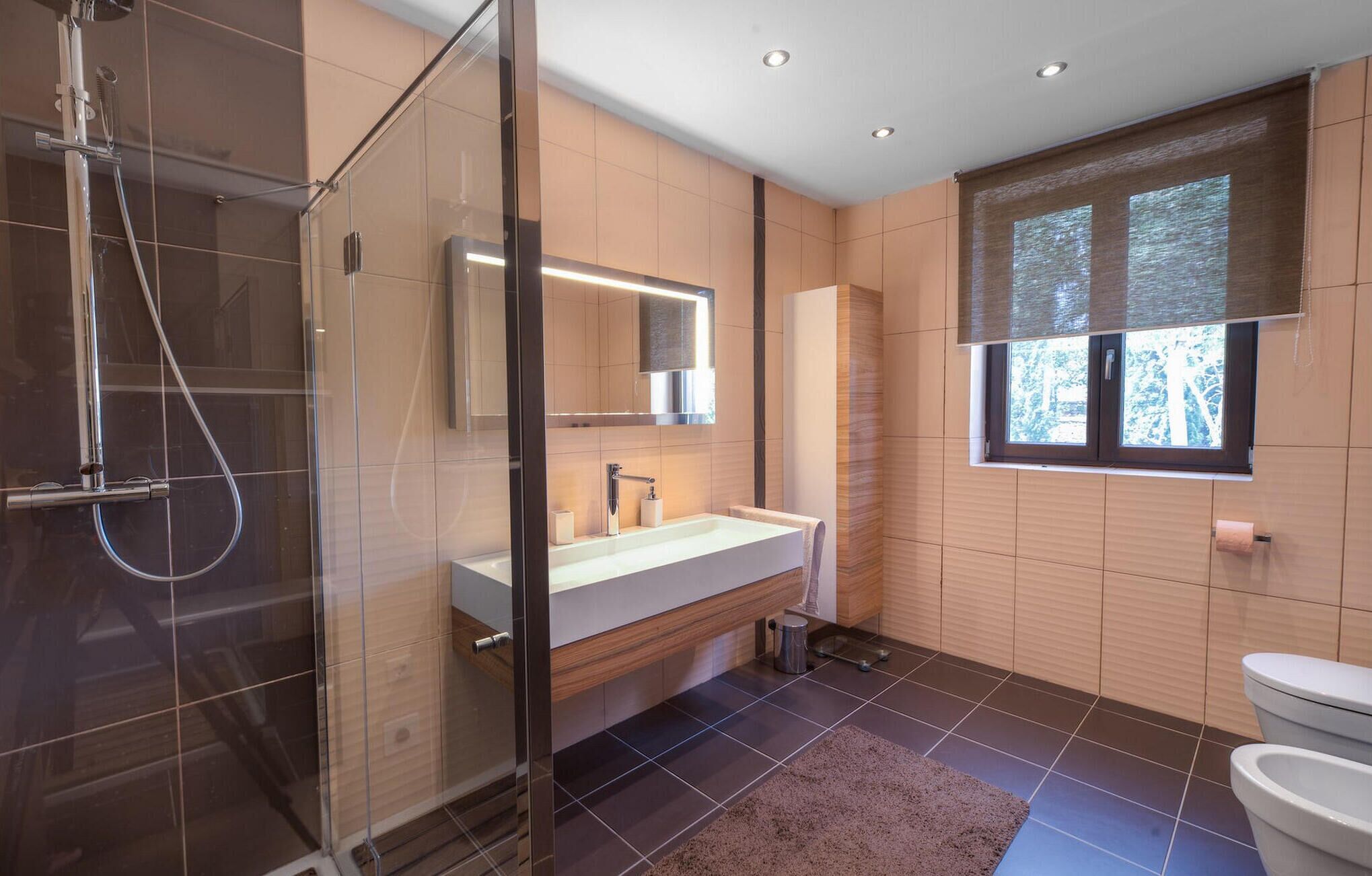 Een moderne badkamer met een douchecabine van glas, een grote spiegel, een wastafel met bijpassend meubel en een raam met uitzicht op bomen.