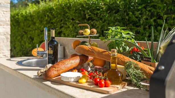 A table with food and drink items, including bread, cheese, tomatoes, and a bottle of wine, set against a backdrop of greenery. - Stunning home in Umag (Umag)