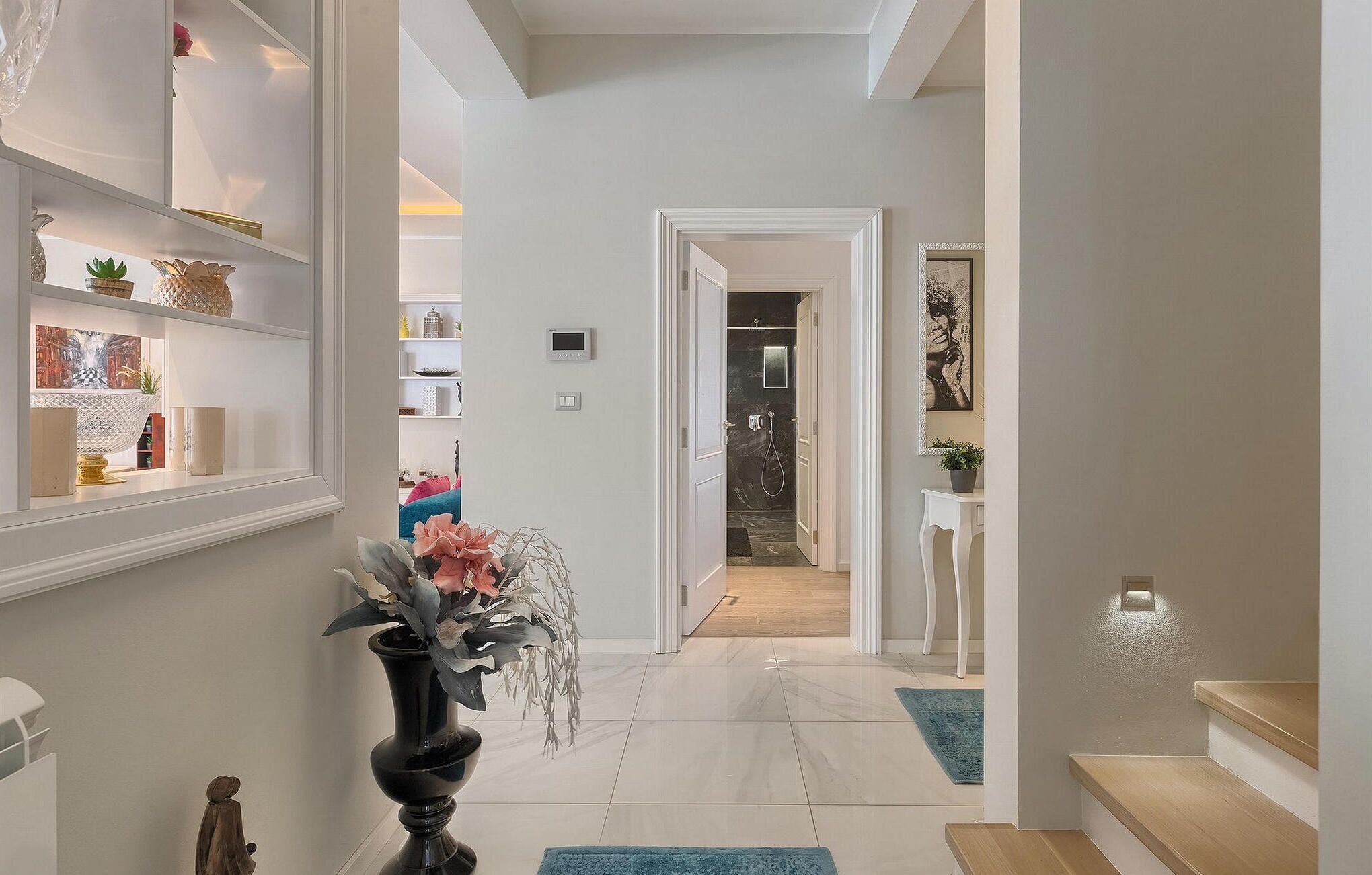 A hallway with a marble floor, white walls, and a staircase. There are shelves with decorative items and a vase with flowers.