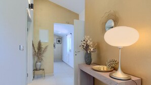 Um corredor com chão claro, um console de madeira com espelho e luminária, e um vaso com flores secas.