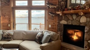 A cozy cabin interior with a stone fireplace, a wooden couch with cushions, and a view of a lake through large windows. - Cozy Cabin on Lake Wisconsin
Skiing, Fishing & Hiking Retreat (Merrimac)