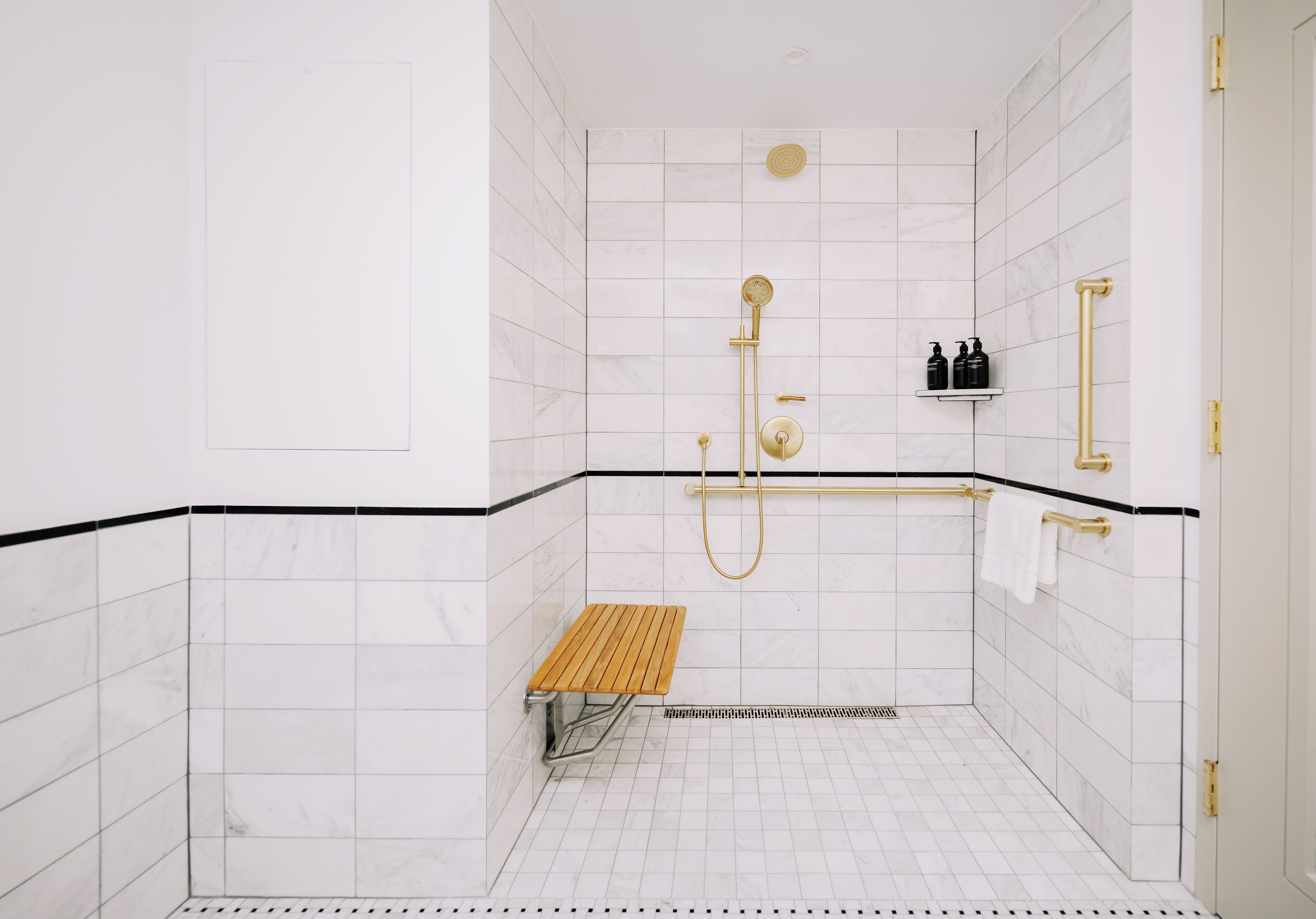 Une petite salle de douche aux murs recouverts de carreaux blancs, avec un banc en bois, des robinetteries dorées et une étagère contenant des articles de toilette.