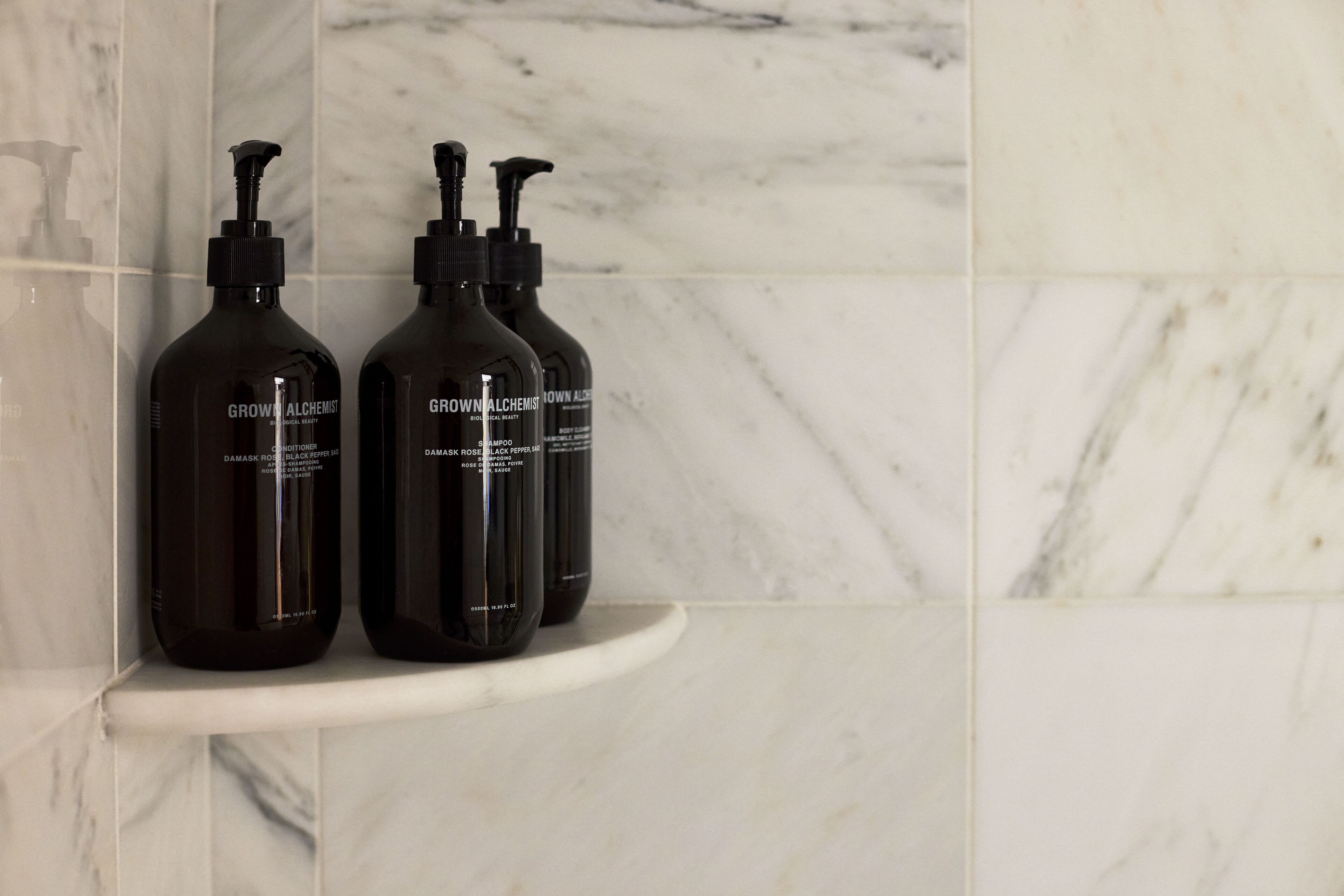 Two bottles of 'Grown Alchemist' shampoo on a white marble shelf in a bathroom.