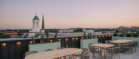 A rooftop terrace with tables and chairs, a view of a cityscape, and a church spire in the background.