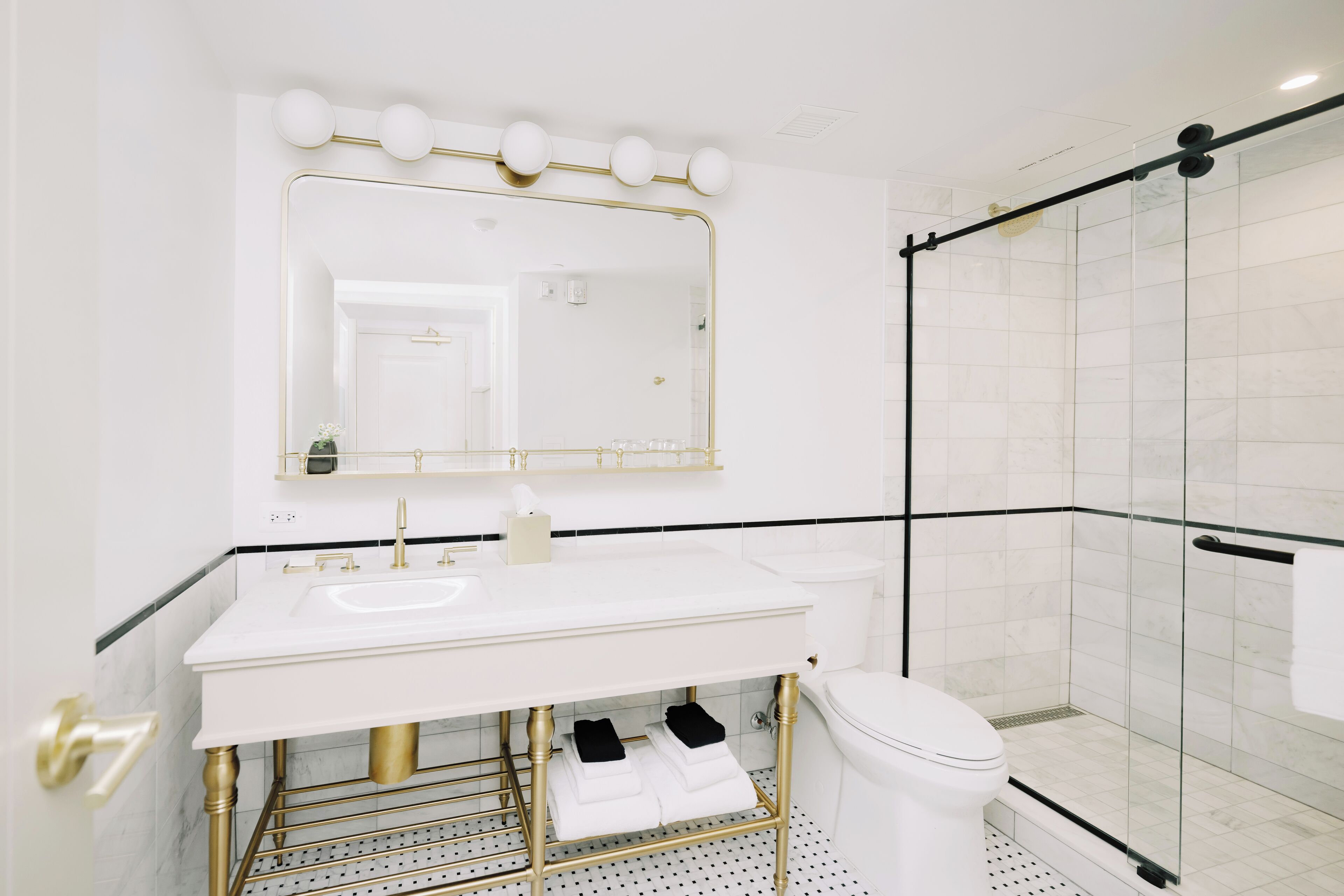 Un baño moderno con lavamanos blanco, detalles dorados, un espejo grande y cabina de ducha con paredes de vidrio.