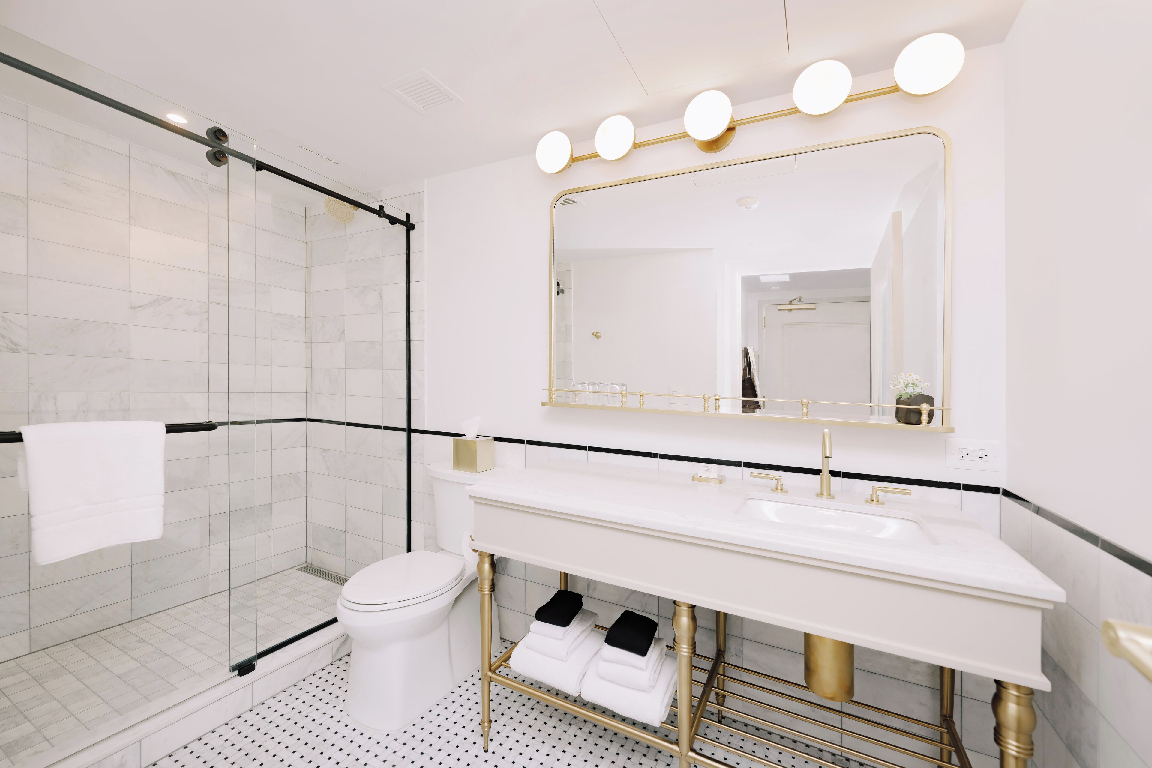 Un baño moderno con cabina de ducha de vidrio, inodoro blanco, tocador con lavamanos dorado y un espejo grande con luces.