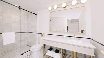 Un baño moderno con cabina de ducha de vidrio, inodoro blanco, tocador con lavamanos dorado y un espejo grande con luces.