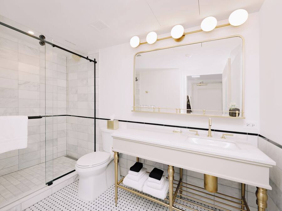Un baño moderno con cabina de ducha de vidrio, inodoro blanco, tocador con lavamanos dorado y un espejo grande con luces.