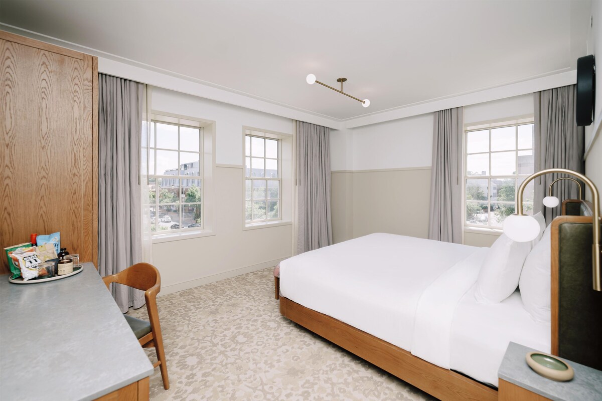 A hotel room with a bed, a desk, a chair, a lamp, and a view of the city.