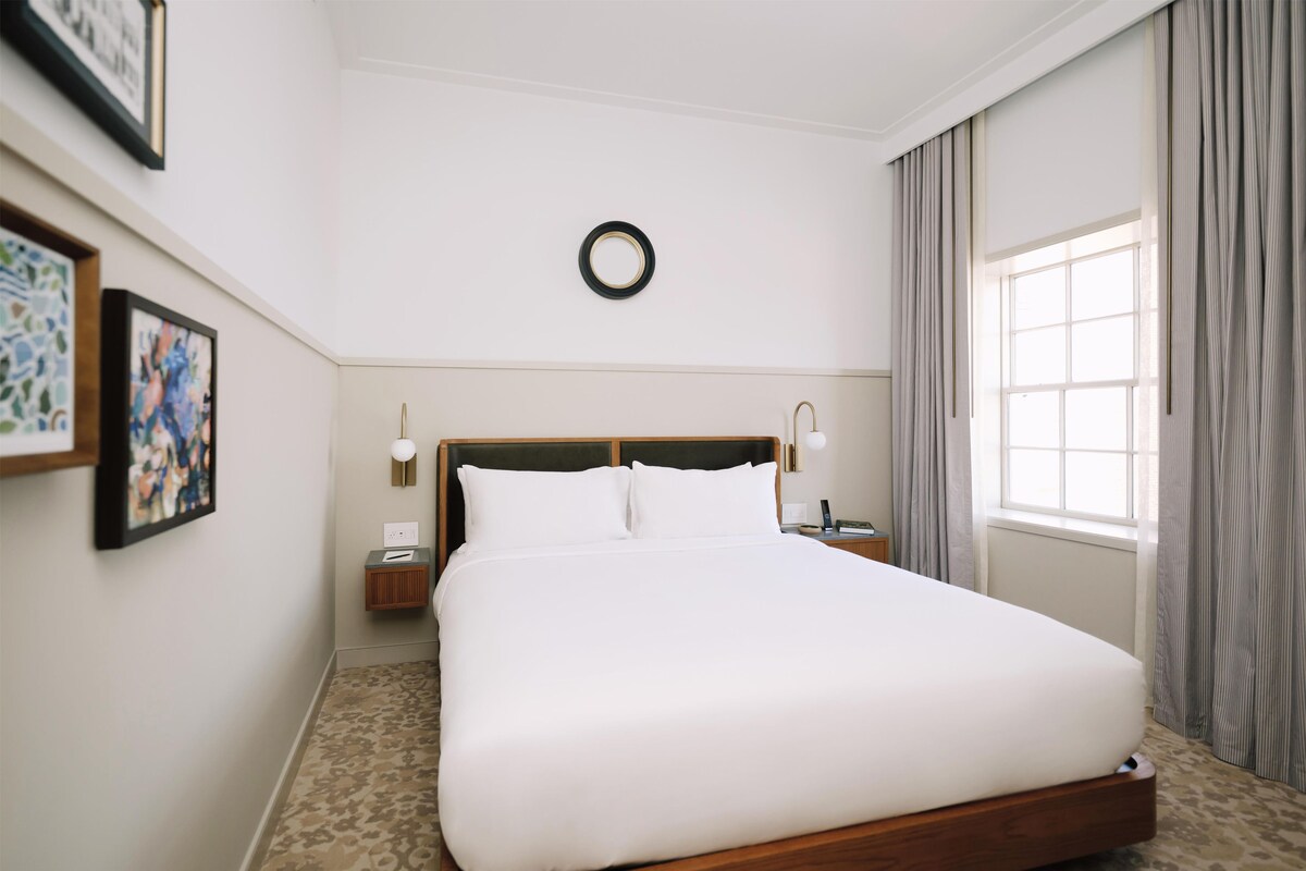 A hotel room with a large bed, a bedside table, a wall clock, and a window with curtains.