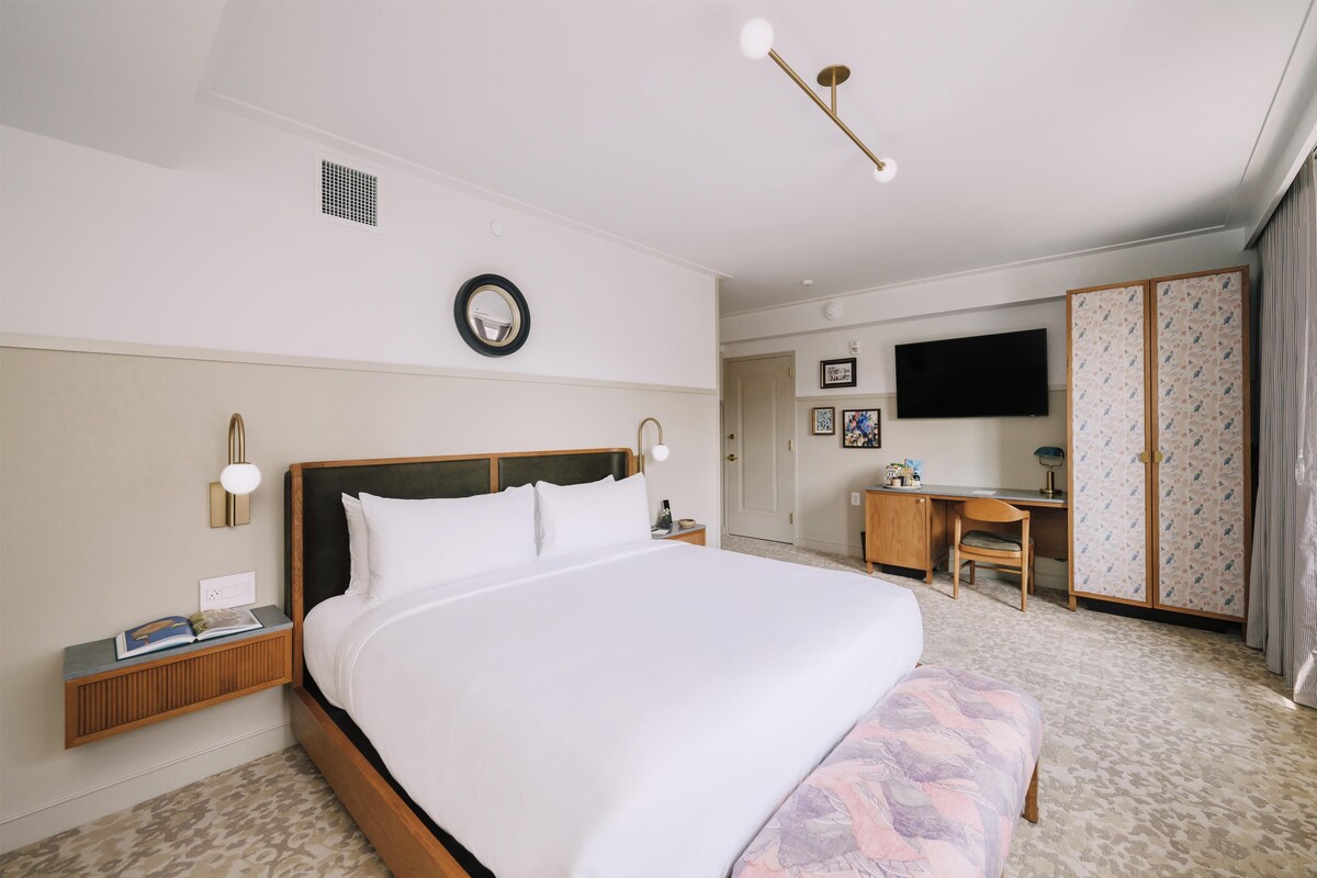 A hotel room with a large bed, bedside table, lamp, wall clock, and a desk with a chair.
