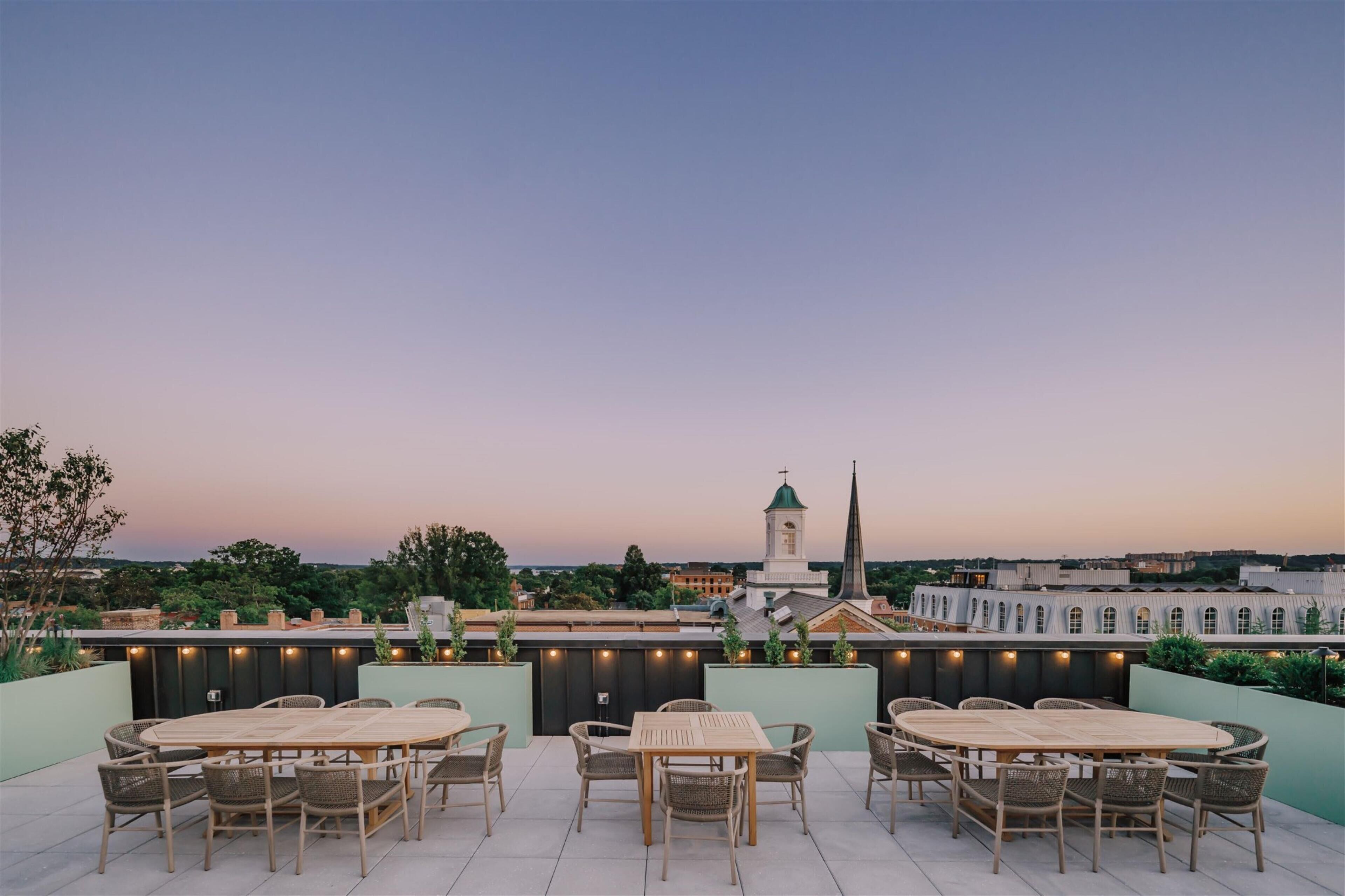 Une terrasse extérieure avec des tables rondes et des chaises en osier, offrant une vue sur le paysage urbain et le clocher d’une église.