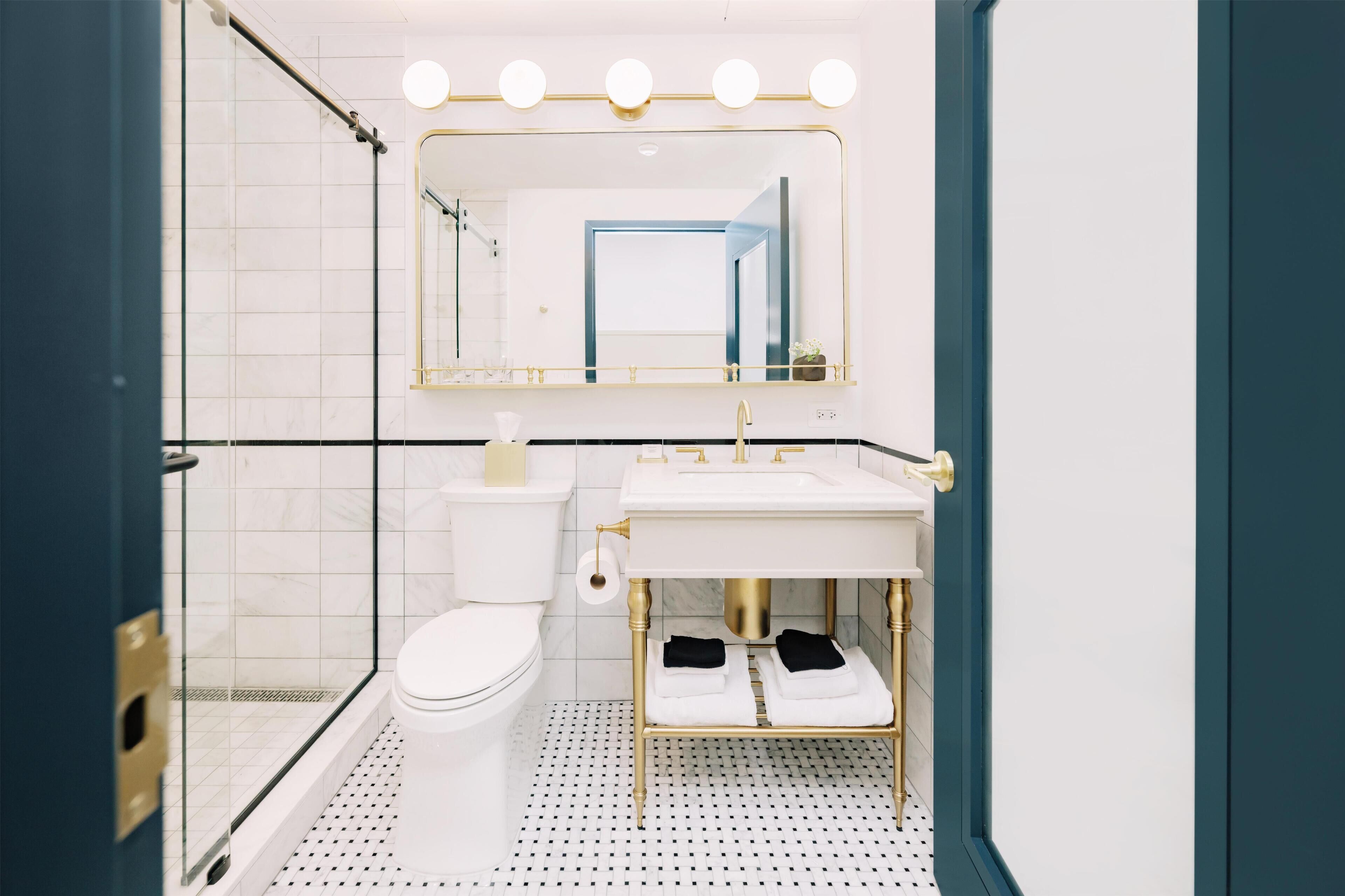 A modern bathroom with a glass shower enclosure, a white toilet, a vanity with a gold sink, and a mirror with lights.