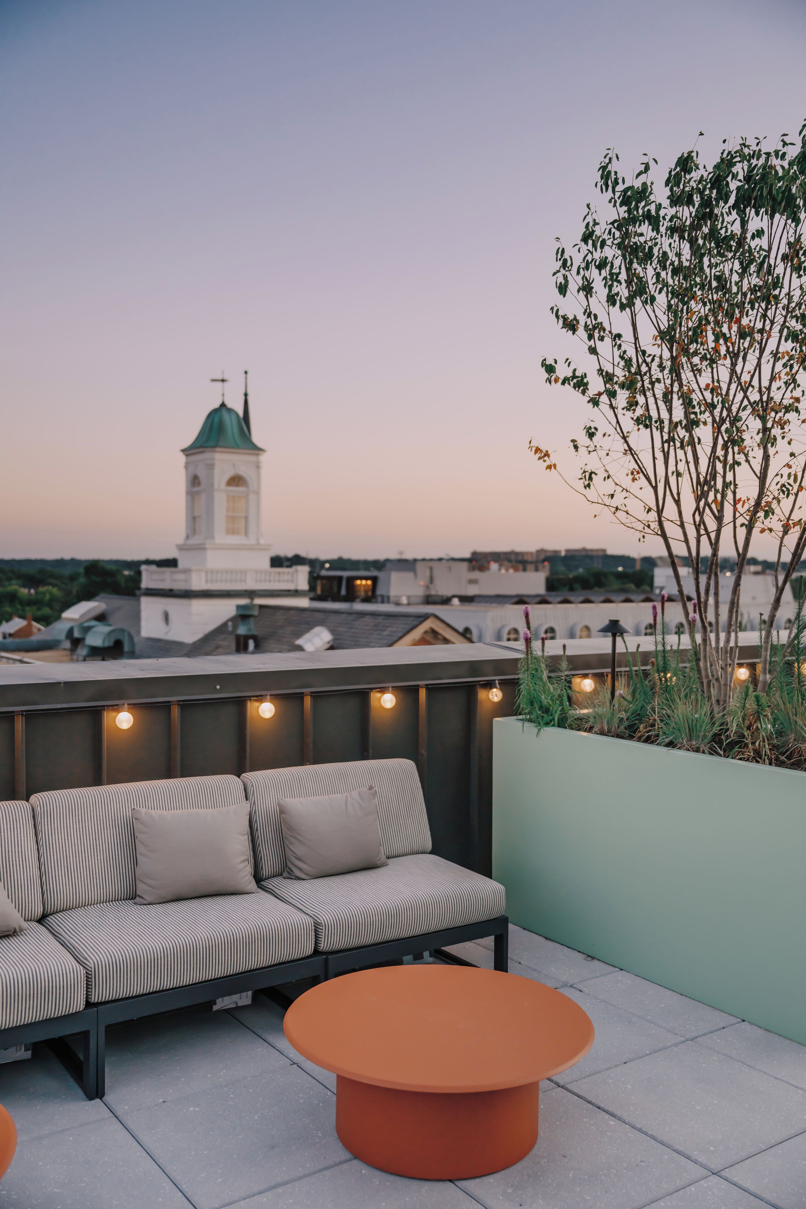 A rooftop terrace with a sofa and round table, a view of a tower, and a green wall with plants.