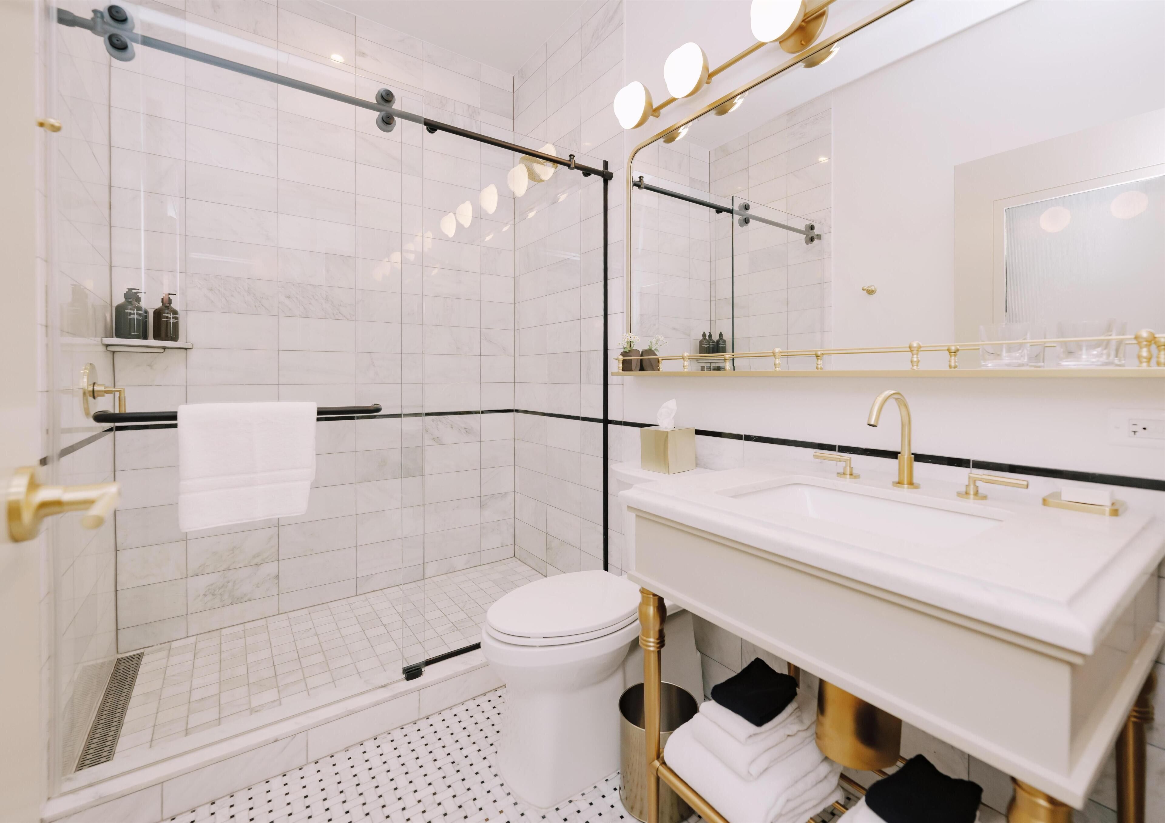 Un baño moderno con cabina de ducha de vidrio, inodoro blanco, tocador con grifo dorado y un espejo grande con iluminación.