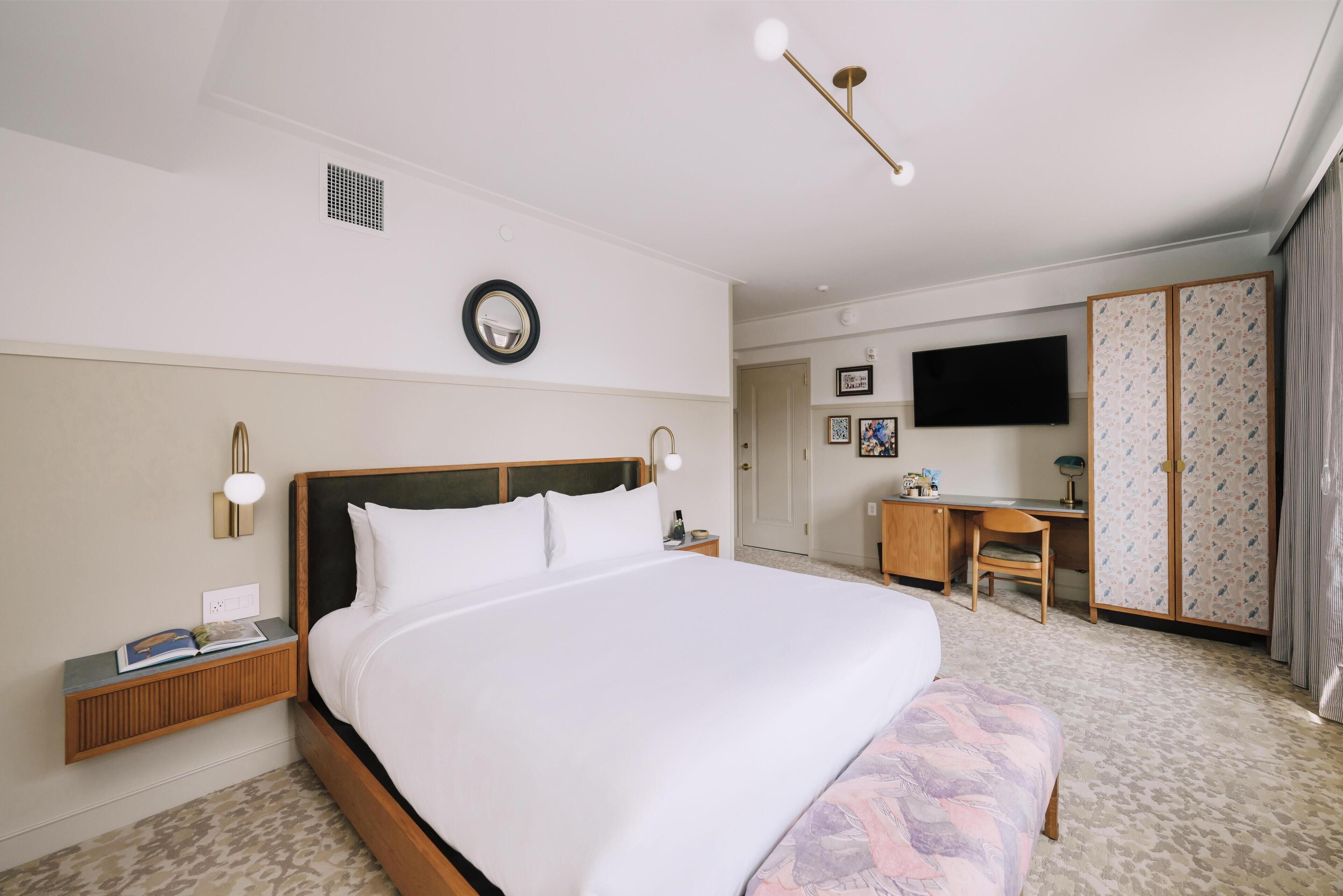 A hotel room with a large bed, bedside table, lamp, wall clock, and a desk with a chair.