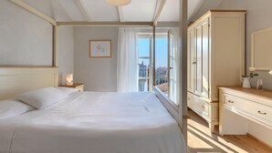 A bedroom with a large bed, a nightstand with a lamp, a dresser, and a window with a view of buildings.
