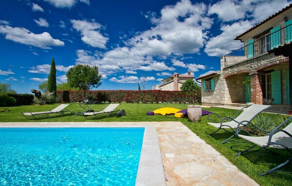 Una piscina amb gandules, un camí de pedra i un edifici de pedra amb persianes verdes.