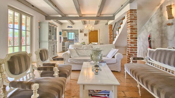 A spacious living room with a high ceiling, exposed beams, and a fireplace. There is a white sofa, a striped armchair, and a coffee table with books. A kitchen is visible in the background. - Pet friendly home in Tinjan (Tinjan)