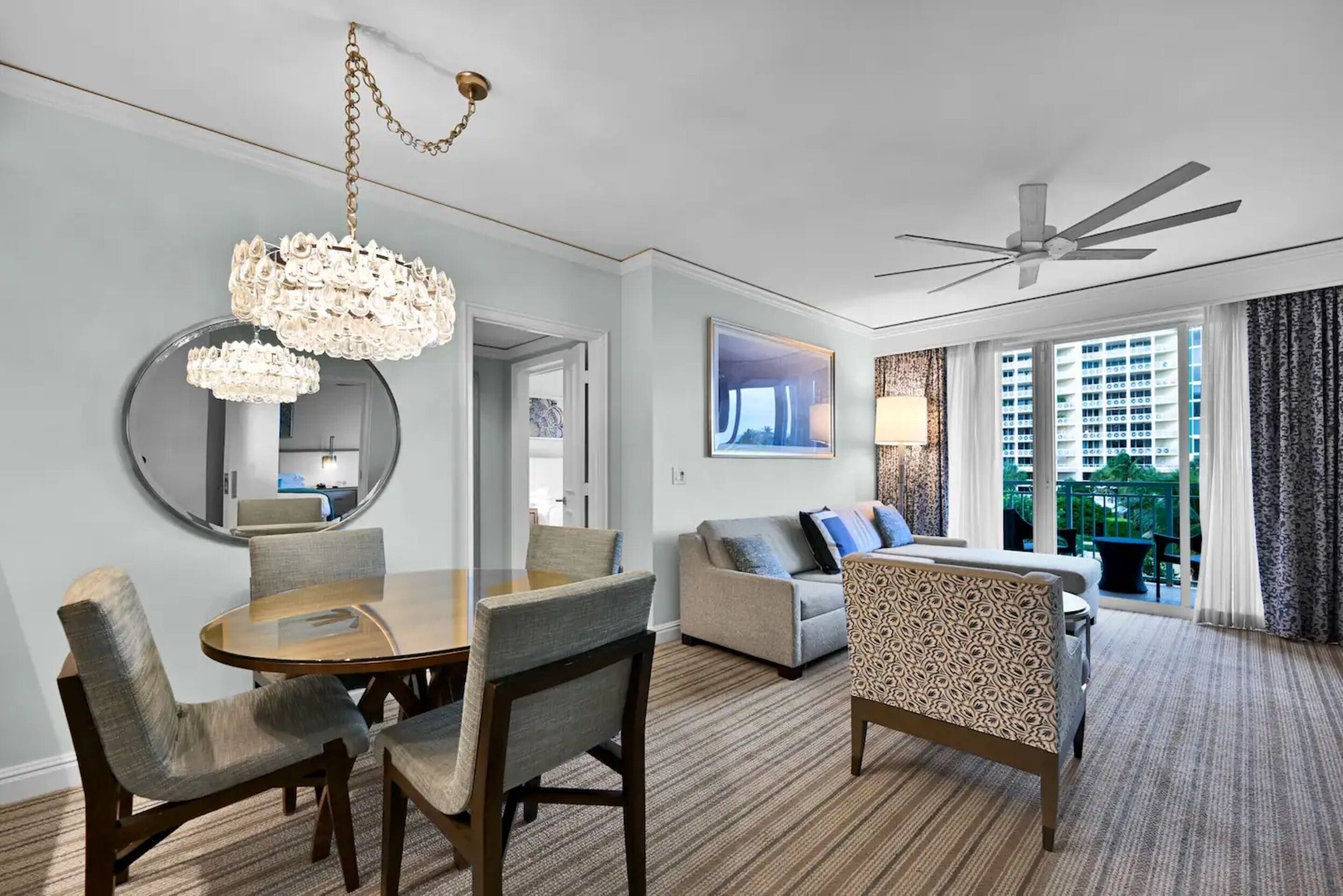 A hotel room with a dining area, a sofa, a patterned armchair, a ceiling fan, and a large window with a view of buildings.