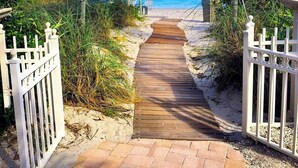 Un sentier en bois mène à une plage, bordée d’une clôture blanche, et un oiseau est perché sur un poteau.