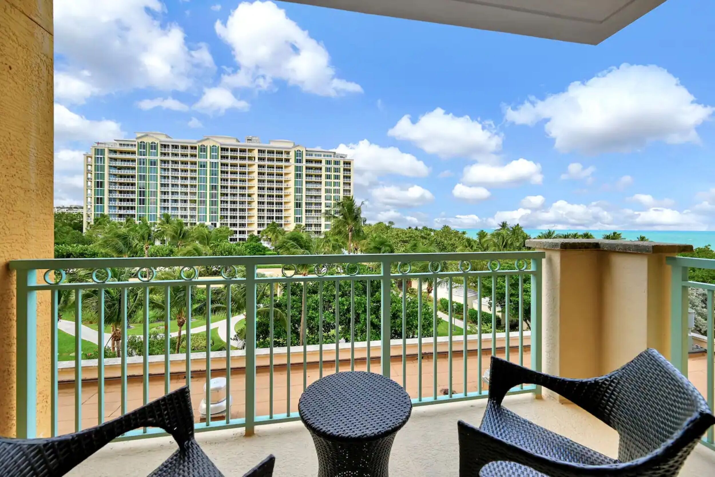 A balcony with wicker chairs and a round table, overlooking a landscaped area with a large hotel building in the background.