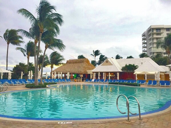 A swimming pool with lounge chairs and palm trees, surrounded by buildings with thatched roofs. - Key Bisyne Suite - Steps to sandy Beaches (Key Biscayne)