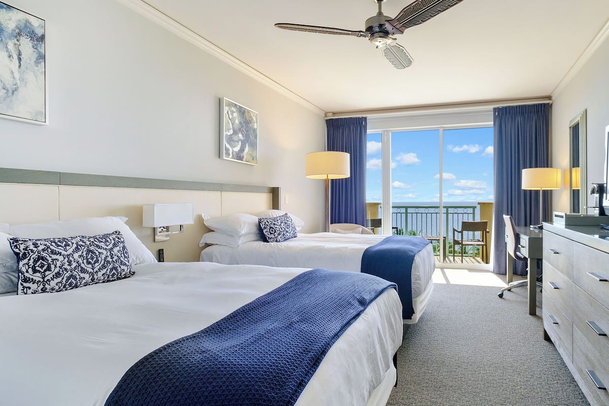 Habitación de hotel con dos camas, un escritorio, televisor y balcón con vistas.