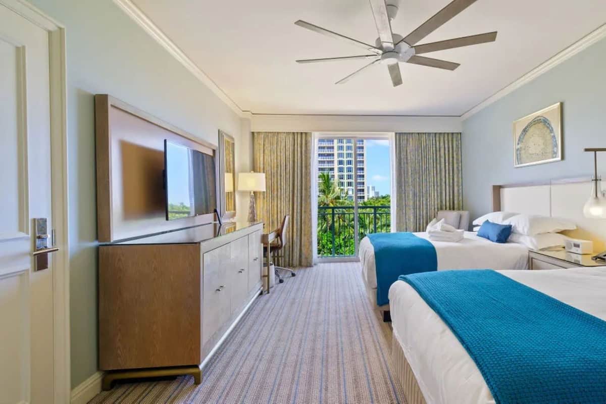 Habitación de hotel con dos camas, ventilador de techo, televisión, escritorio y balcón con vistas a la ciudad.