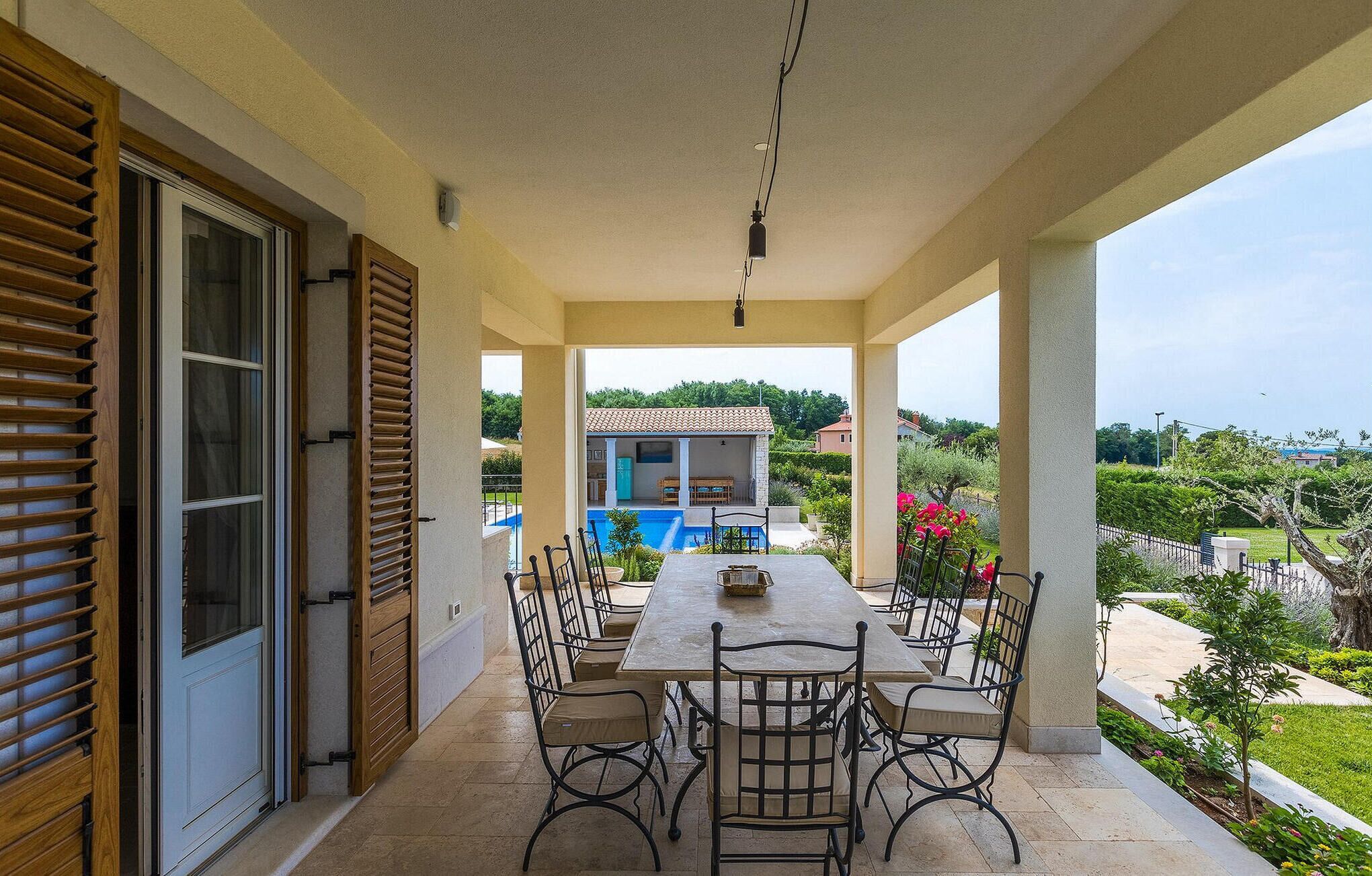 Eine überdachte Terrasse mit Tisch und Stühlen, Blick auf einen Swimmingpool und ein Gebäude im Hintergrund.